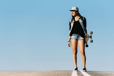 Woman holding skateboard standing on road against clear blue sky