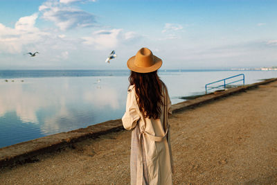Back view of woman in coat and hat standing on pier , seagulls flying in blue sky.