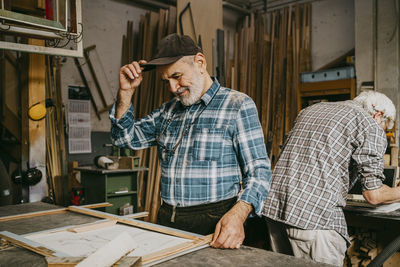 Senior male carpenter adjusting cap while working with colleague at repair shop