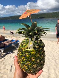 Cropped hand holding pineapple at beach against sky