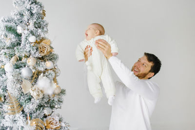 A young happy dad lifts up a baby in his arms near the christmas tree. happy fatherhood
