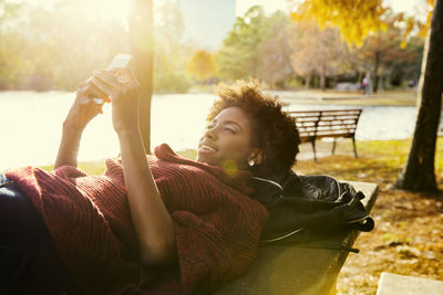 Young woman using phone while lying on bench at park