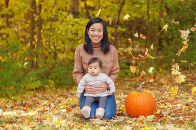 Portrait of smiling mother holding cute daughter while kneeling on autumn leaves in park
