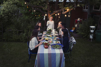 Happy woman standing at dining table with friends enjoying dinner party at yard