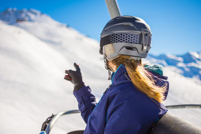 Woman wearing ski goggles and helmet while sitting in ski lift against sky