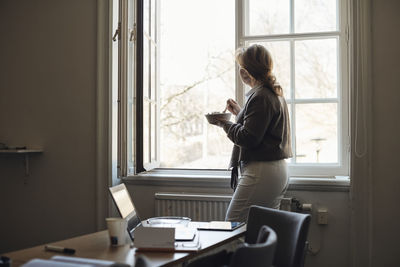 Side view of teacher eating lunch while looking outside window in classroom