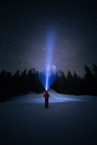 Rear view of person holding illuminated flashlight while standing on snow covered field against sky at night