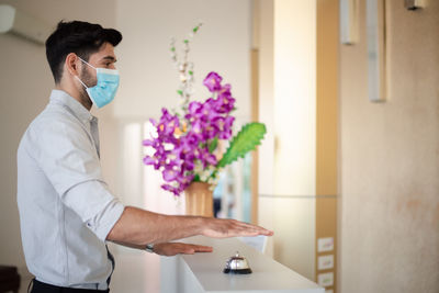 Side view of young businessman wearing mask pressing service bell at hotel reception