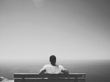 Rear view of man sitting on bench by sea against clear sky