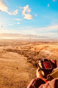 Cropped hands of man holding camera on landscape against sky during sunny day