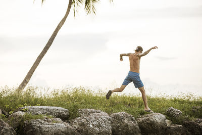 A young man without shirt jumps on rocks at near palm tree at water