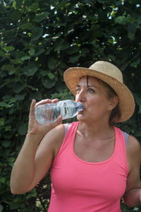 Mature woman drinking water while standing against plants