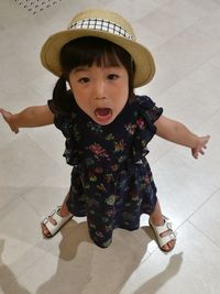 High angle portrait of cute girl with arms outstretched wearing hat while standing on floor