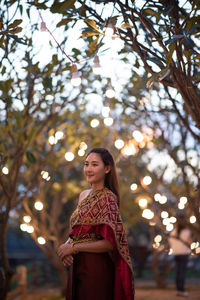 Thoughtful woman wearing traditional clothing while standing against illuminated trees during sunset