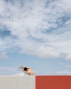 Rear view of shirtless young man with arms raised standing on building terrace against cloudy sky