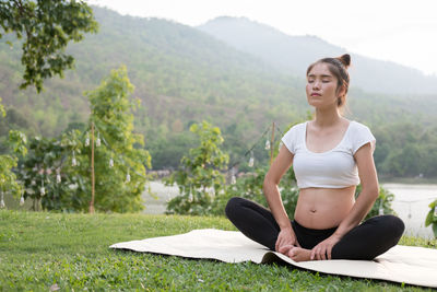 Pregnant woman with arms raised exercising while sitting on mat over grass against sky