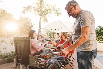 Man cooking food on barbecue grill while family enjoying meal in background at yard