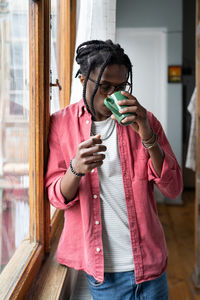 Upset african guy standing near window holding mug drinking tea to calm down and reduce stress