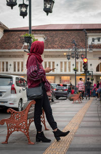 Side view full length of woman standing by bench on sidewalk in city