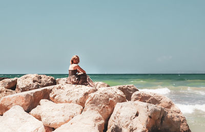 Side view of mid adult woman sitting on rock by sea against clear sky during sunny day