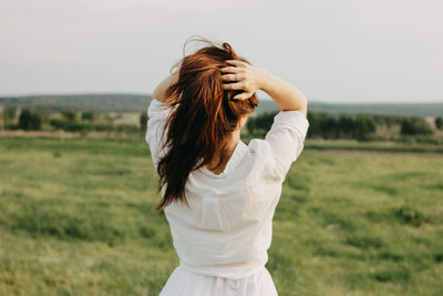 Rear view of woman with hands in hair standing on grassy land against clear sky