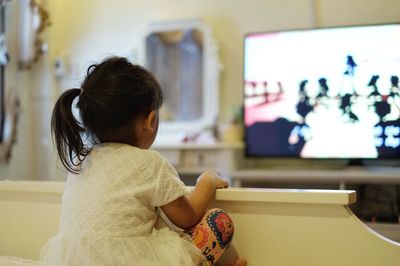 Rear view of girl watching tv while sitting on bed at home