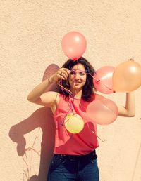 Smiling young woman holding balloons against wall during sunny day