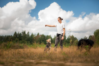 Man playing with dogs while standing on grassy land against cloudy sky