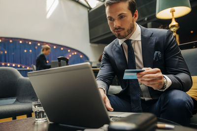 Young businessman holding credit card using laptop while sitting in hotel lounge
