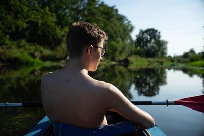 Midsection of shirtless man sitting by lake against trees