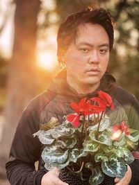 Portrait of young man holding red flowering cyclamen plant against trees and setting sun