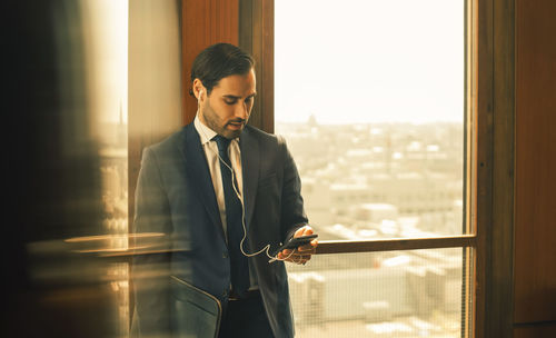 Mid adult legal professional using smart phone while standing against window at law office