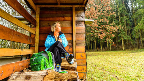 Woman looking away while sitting against log cabin in forest