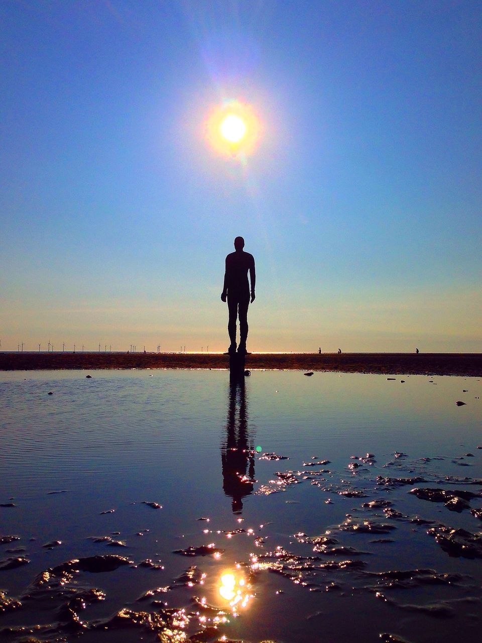 water, sun, sunset, reflection, silhouette, lifestyles, leisure activity, standing, full length, sea, sunlight, tranquility, tranquil scene, beach, scenics, clear sky, lens flare, sky