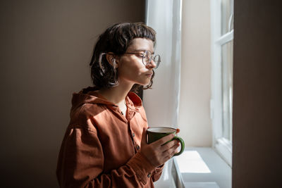 Teen girl nerd in glasses looking at window drinking cup of tea at home. introvert, friendless