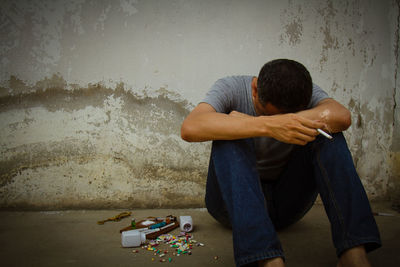 Addicted man sitting with cigarette and pills against wall