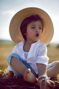 Baby boy in straw hat and blue pants sitting on a haystack in a field in autumn