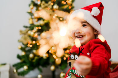 Cute girl holding sparkler while sitting against christmas tree