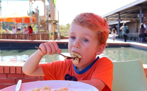 The blond boy eats pasta and looks intently into the distance. a   boy eats lunch at a street café.