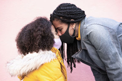 Mother and daughter wearing face mask standing face to face against pink wall