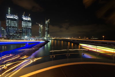 Light trails on river by illuminated buildings against sky at night