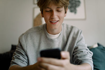 Smiling teenage boy using smart phone while sitting at home