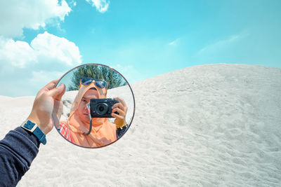 Reflection of woman photographing with camera on mirror at beach against blue sky