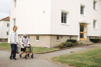 Female home caregiver walking with elderly woman by building on footpath