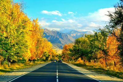 Road amidst trees against sky leading towards mountains during autumn