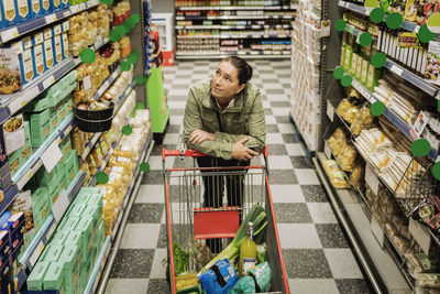 High angle view of thoughtful woman leaning on shopping cart at supermarket