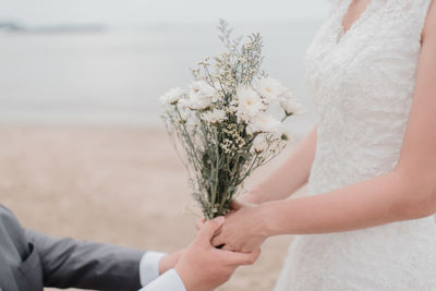 Midsection of groom holding hands of bride with bouquet at beach
