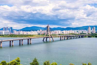 Bridge over river with city in background against cloudy sky