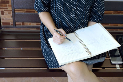Midsection of female student writing on book while sitting on bench