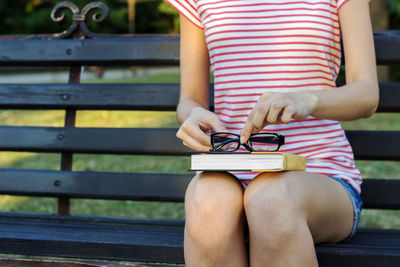 Midsection of woman with book and eyeglasses sitting on bench at park